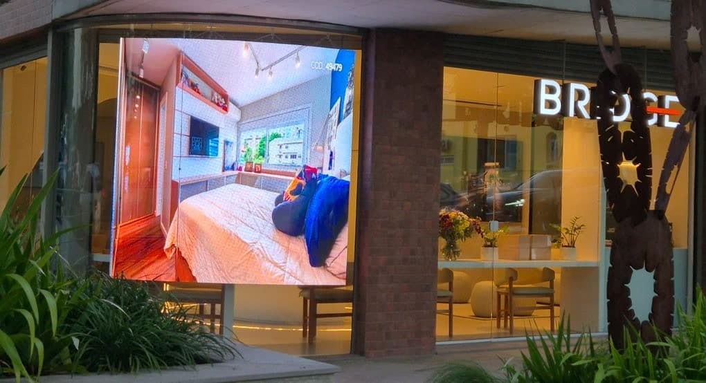 Vitrine curva de vidro com painel de LED integrado à fachada da Bridge, vegetação e escultura em primeiro plano.