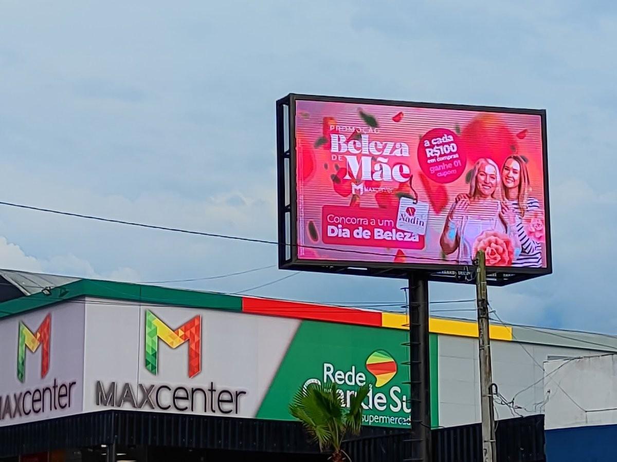 Totem de LED comercial em operação junto à loja Maxcenter, exibindo campanha promocional, fachada da loja visível ao fundo.