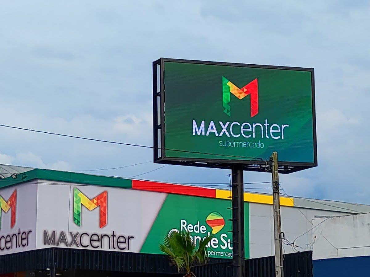 Painel de LED em totem sobre poste, junto à fachada de outra loja MaxCenter, exibindo identidade da marca.
