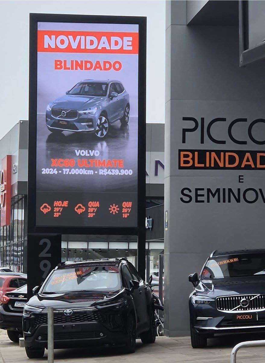 Totem de LED da Piccoli Blindados exibindo anúncio de veículo do estoque e previsão do tempo, veículos blindados em primeiro plano.
