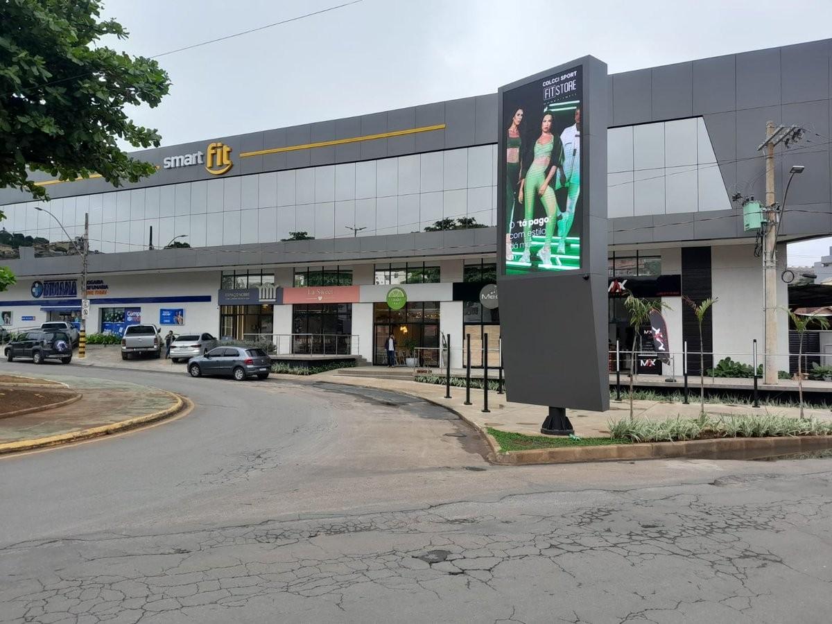 Totem de LED junto a fachada comercial ativa, rua com tráfego de veículos e pedestres.